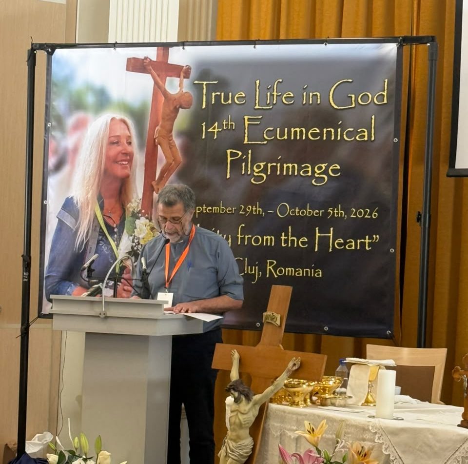 El Padre Carlos White de Argentina hablando en un podio frente a una pancarta del 14° Peregrinaje Ecuménico 'Vida Verdadera en Dios', con una cruz en el fondo. La pancarta incluye detalles del evento y una foto de Vassula sosteniendo flores.