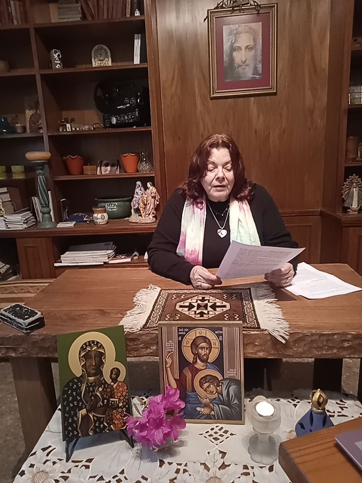 Una mujer sentada en un escritorio de madera, leyendo un documento. En la mesa hay íconos religiosos, una flor y una vela encendida. Al fondo, una imagen de Jesús en la pared y estantes con objetos decorativos.